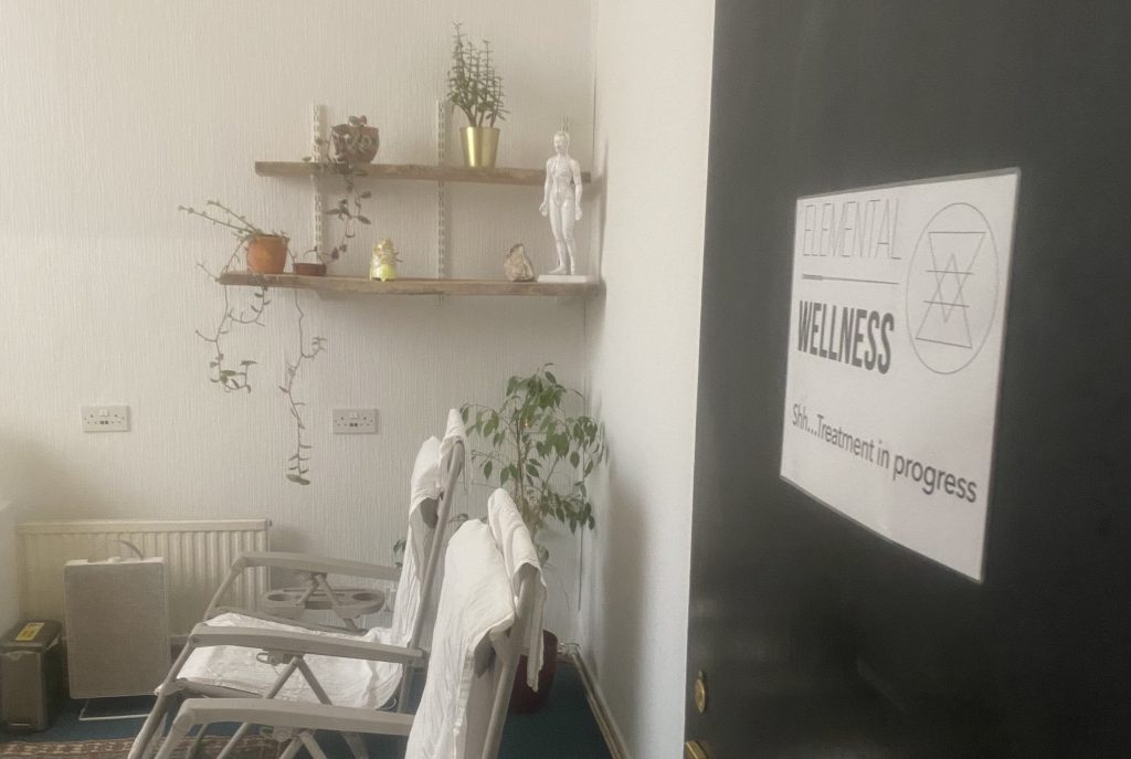 Group treatment room with white walls, wooden shelves holding potted plants and decor, and row of white reclining chairs covered with towels